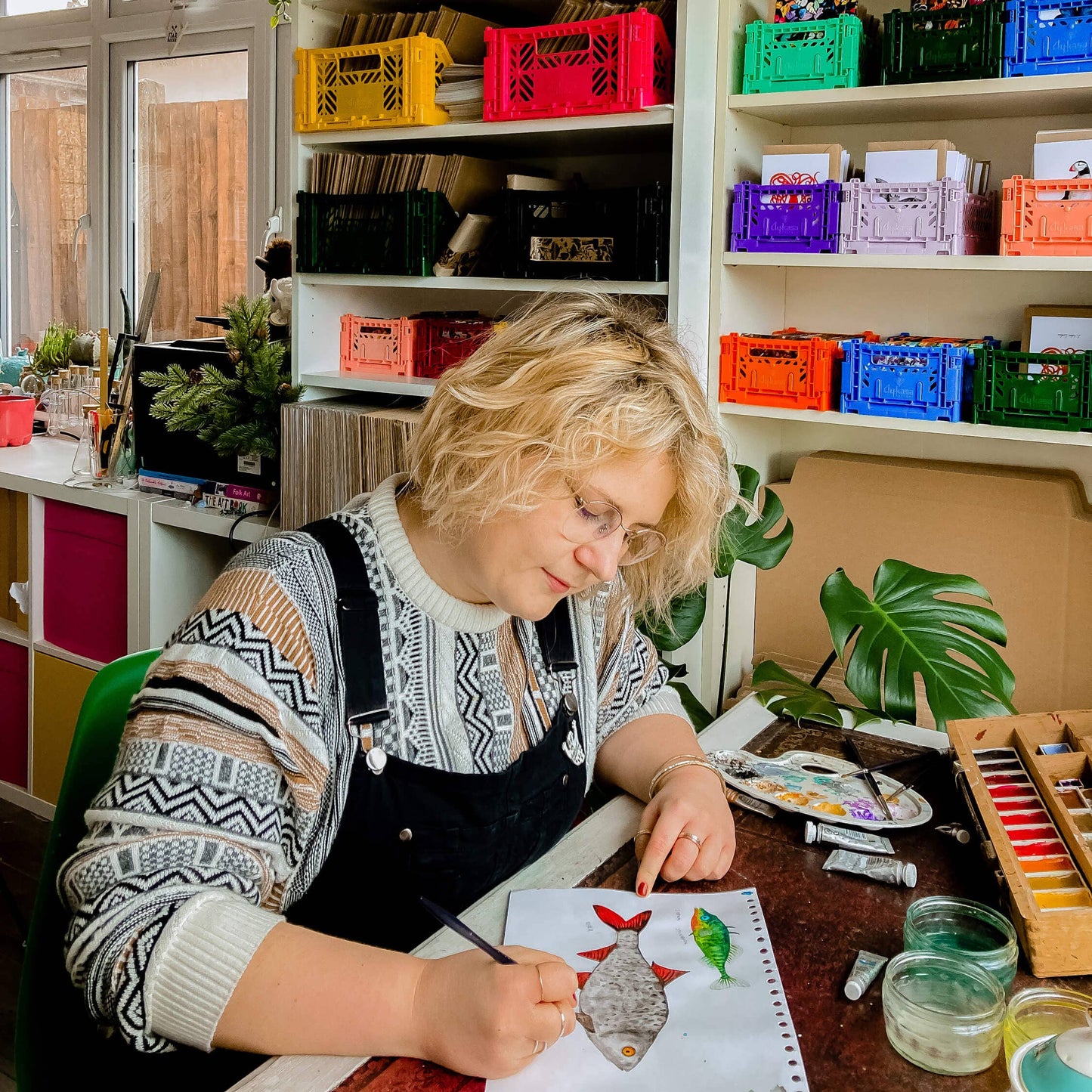 Person painting at a desk with colorful crates on shelves in the background