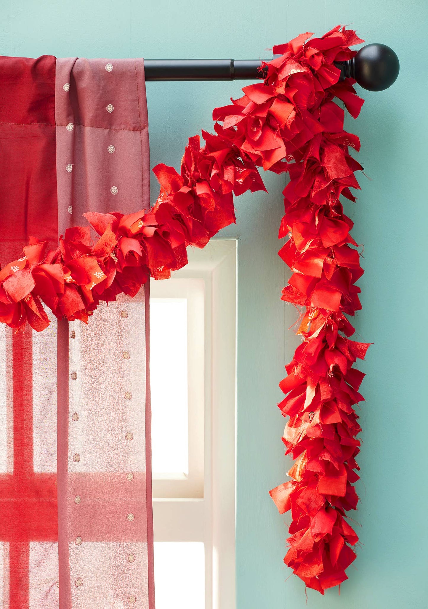 Red ruffled sari garland draped over a black curtain rod against a light blue wall.