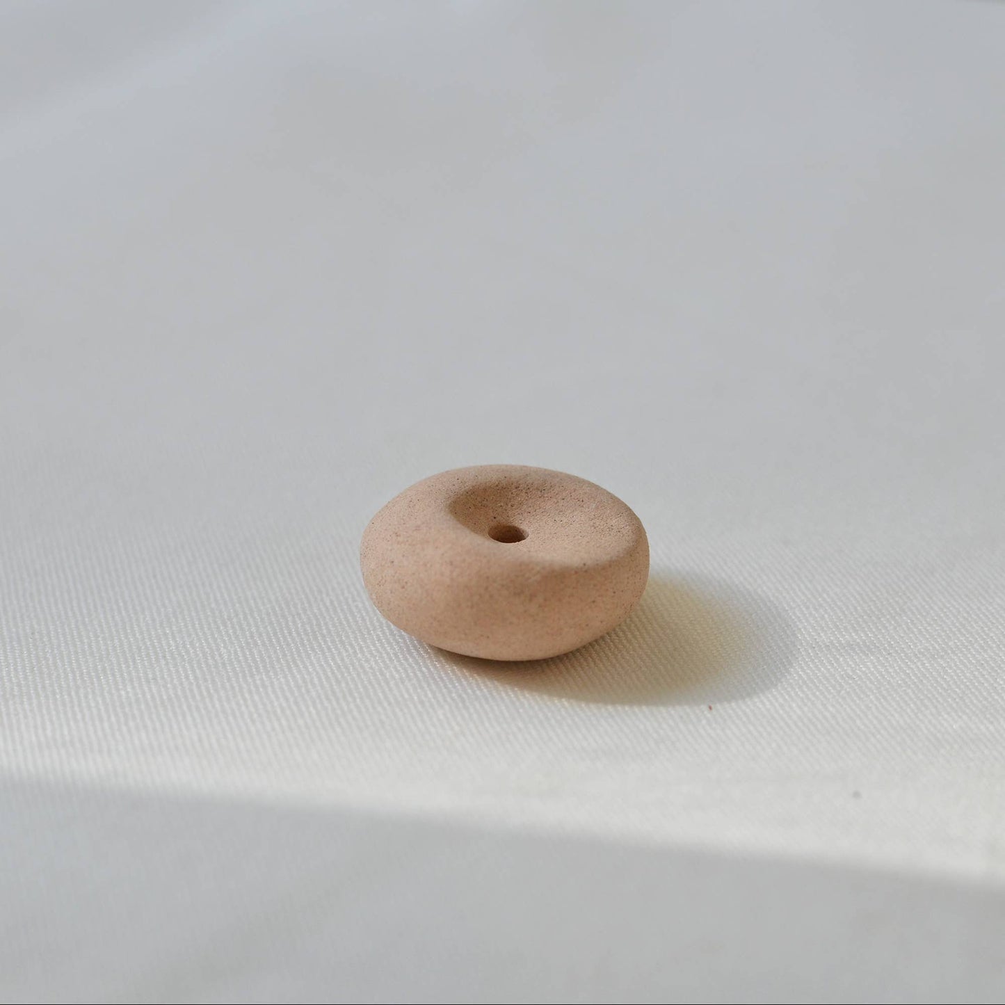 Clay pebble incense holder in soft beige tones, shown on a pale fabric background with a raw, organic finish.