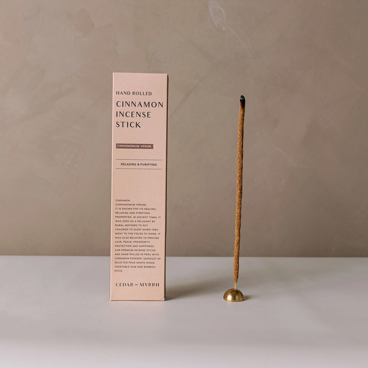 A hand rolled cinnamon incense stick presented next to its packaging, which has a minimalistic design with text.