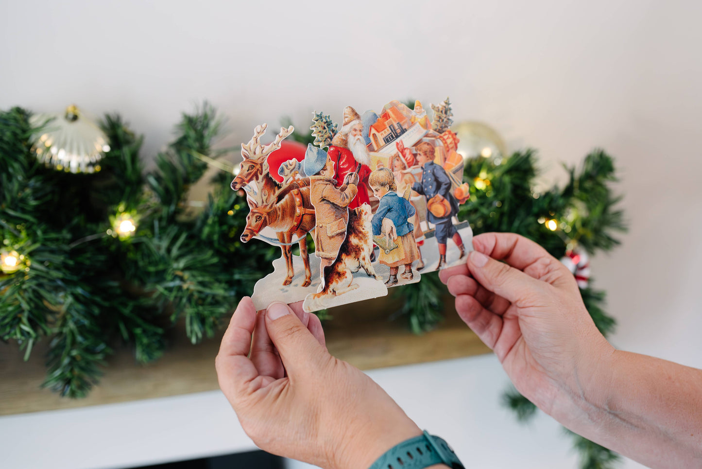 Person holding a Christmas-themed paper cutout with Santa Claus and reindeer in front of a decorated Christmas tree.