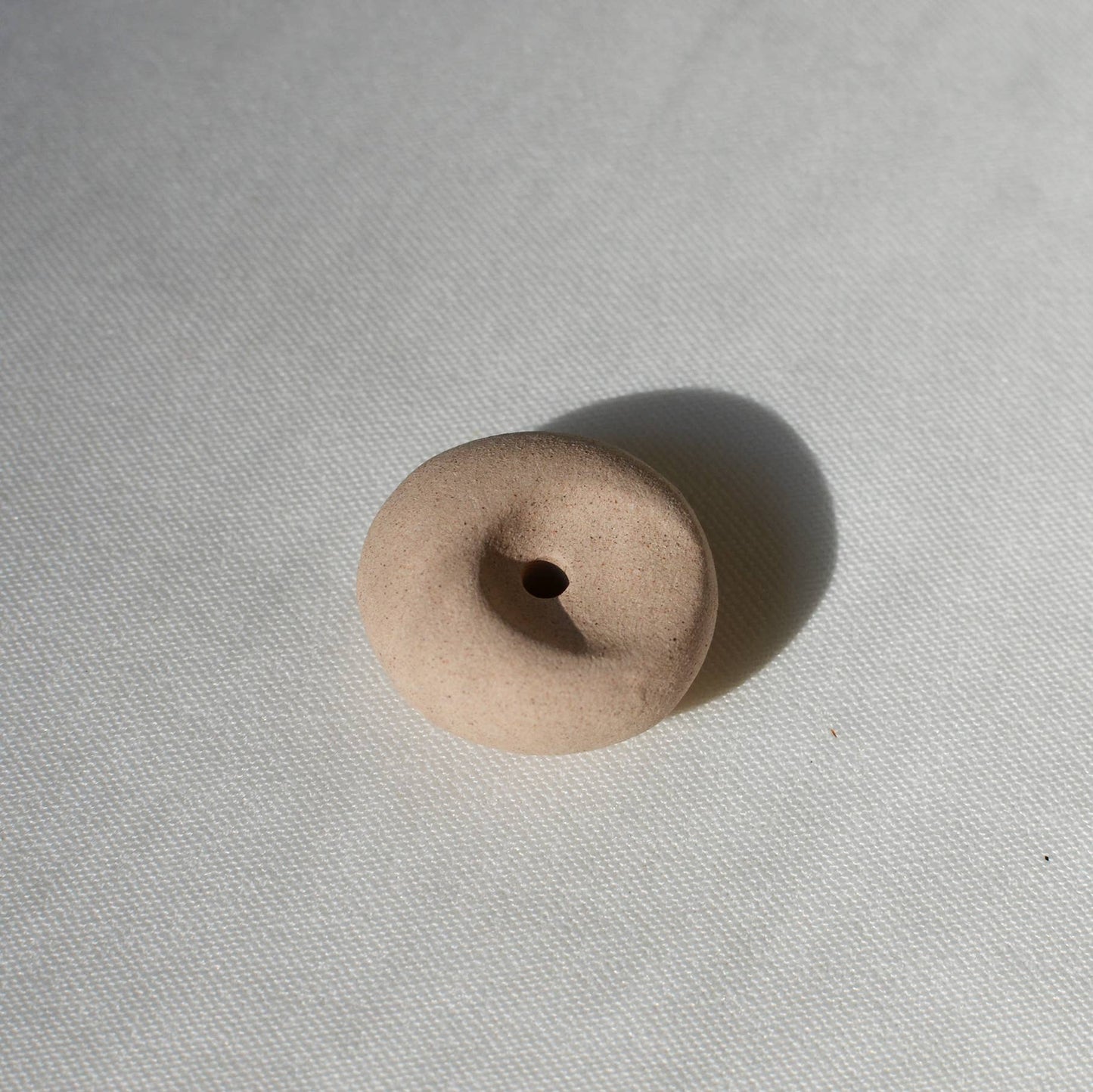 Clay pebble incense holder in soft beige tones, shown on a pale fabric background with a raw, organic finish.