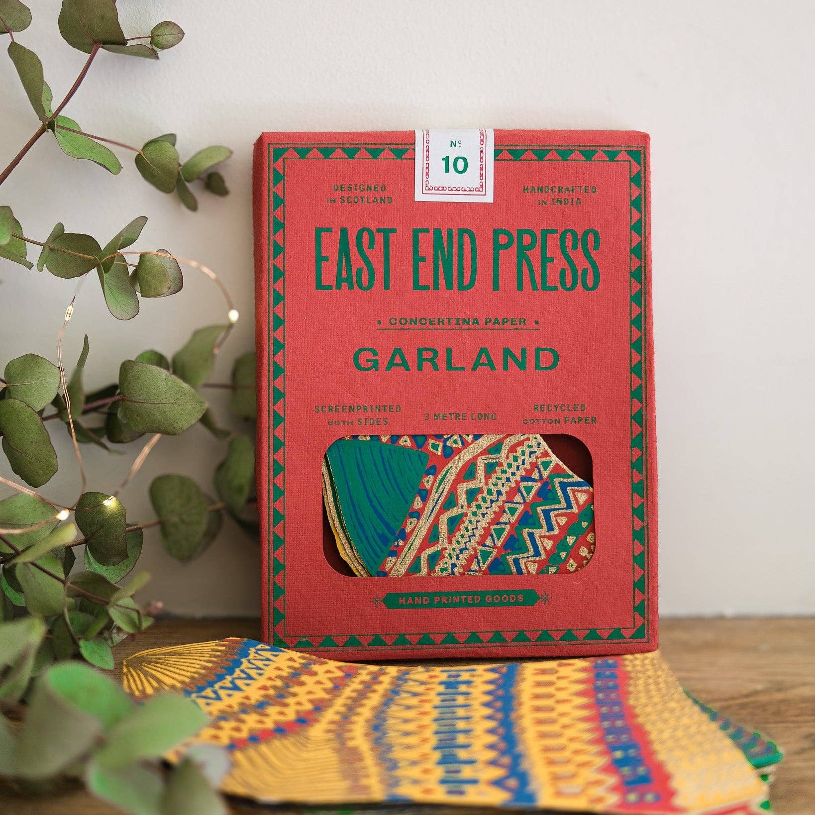 Red box labeled 'East End Press Garland' with colorful packaging on a wooden surface next to greenery.
