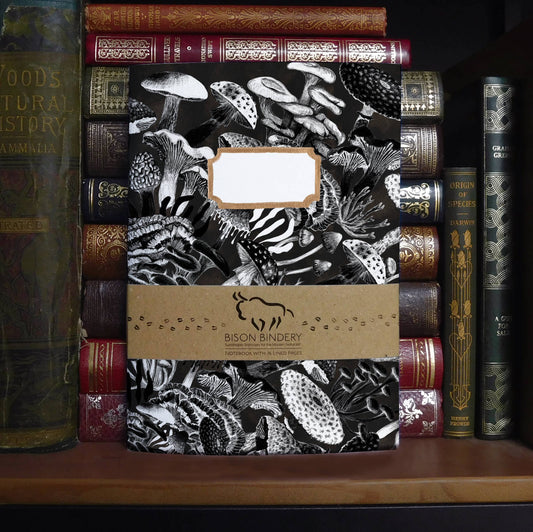 recycled paper notebook with black and white mushroom design and brown band, placed on a shelf with antique books.
