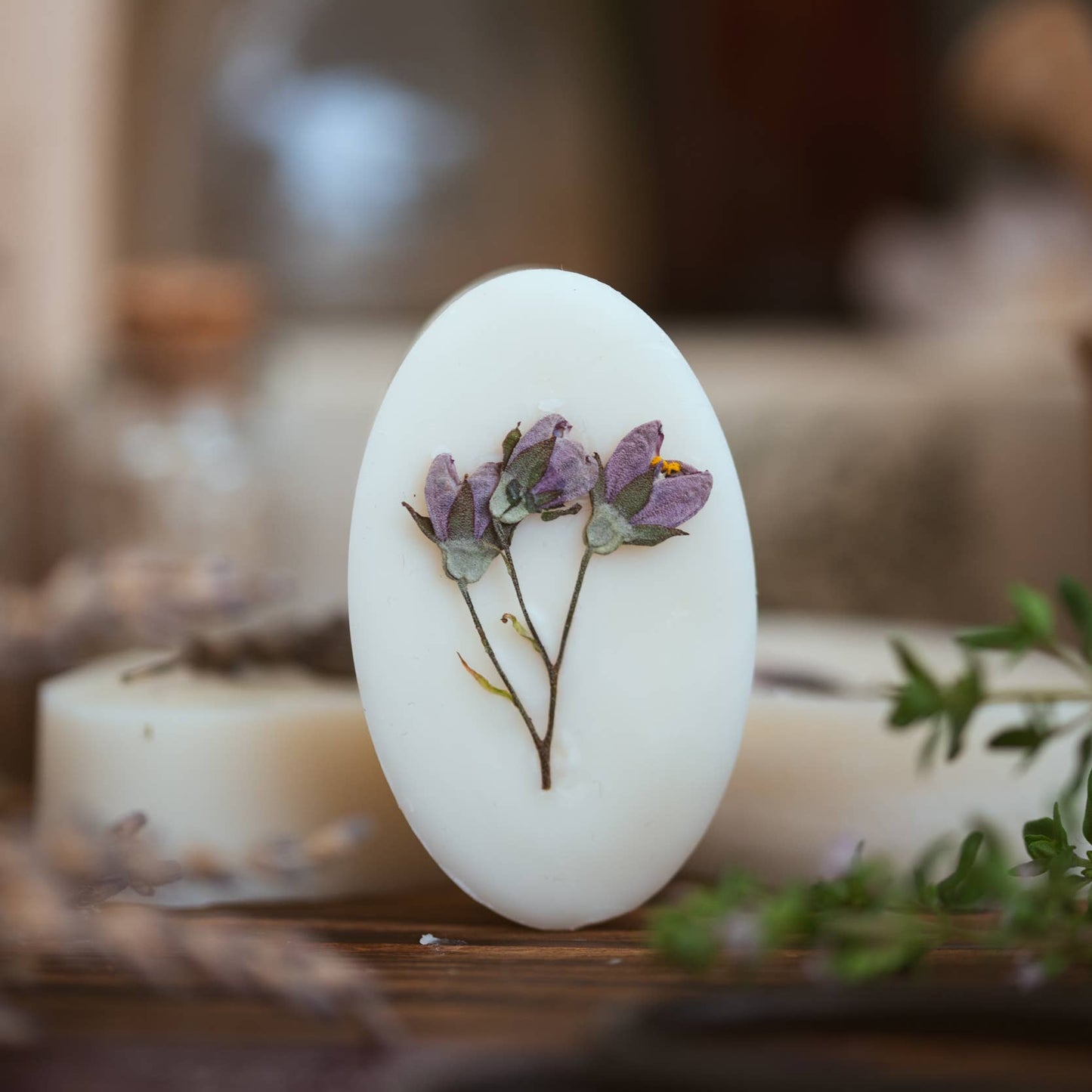 An oval white wax melt with a pressed purple flower and green stem, standing upright on a wooden surface with blurred candles and bottles in the background.