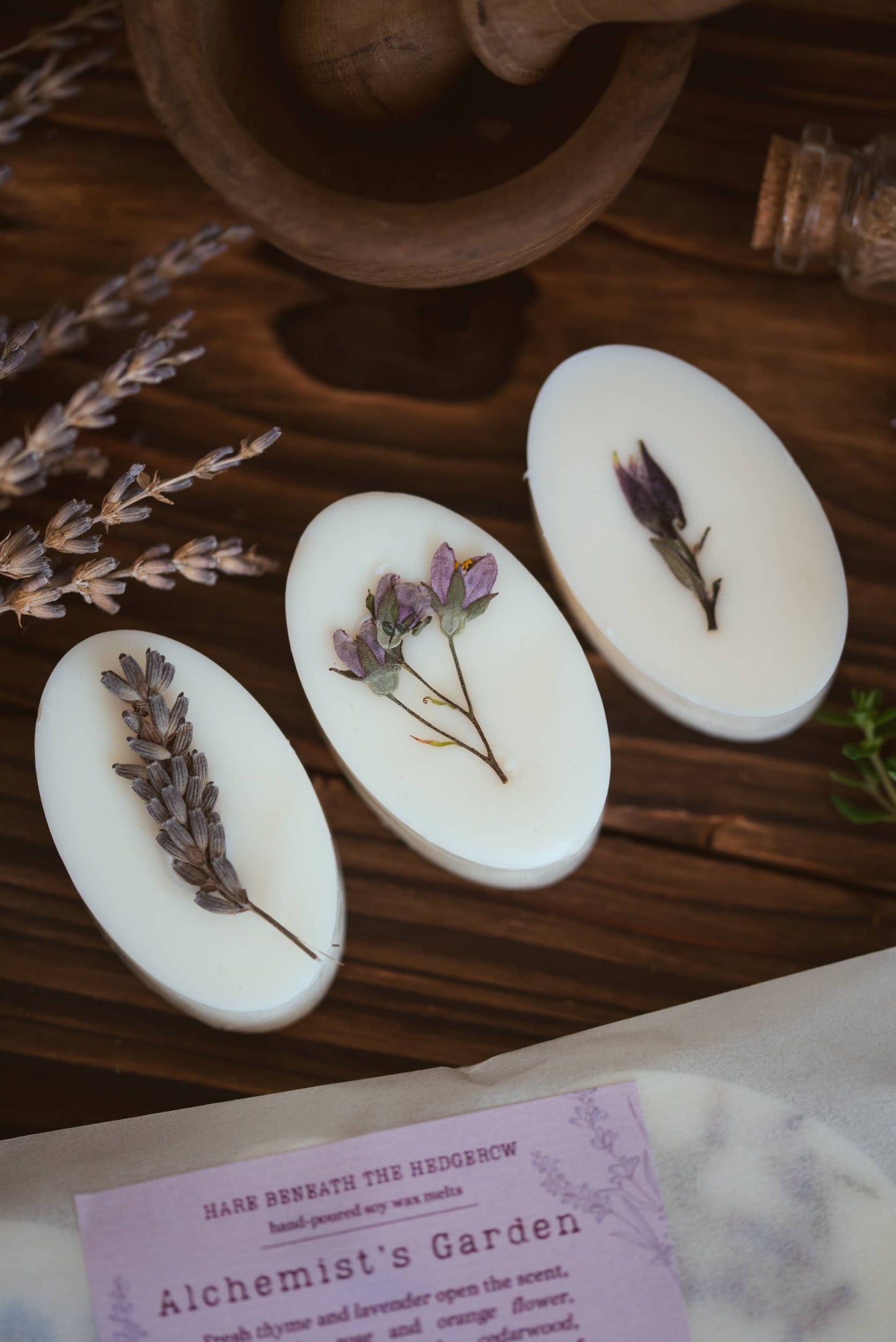 Three oval white wax melts with pressed dried botanicals are arranged on a dark wooden surface next to lavender sprigs and a branded card reading 'Alchemist's Garden'.