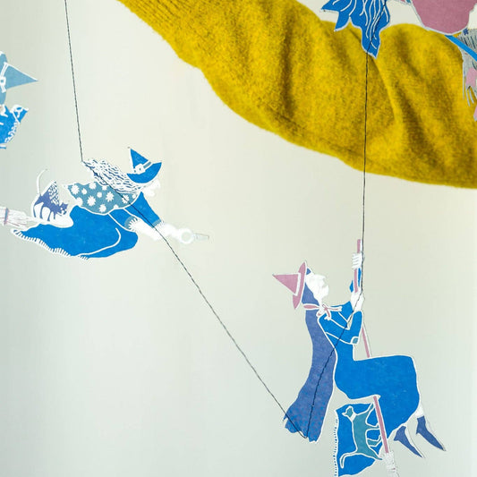 Colorful paper cutout witches mobile with blue and pink animals held by a person wearing a mustard yellow sweater.