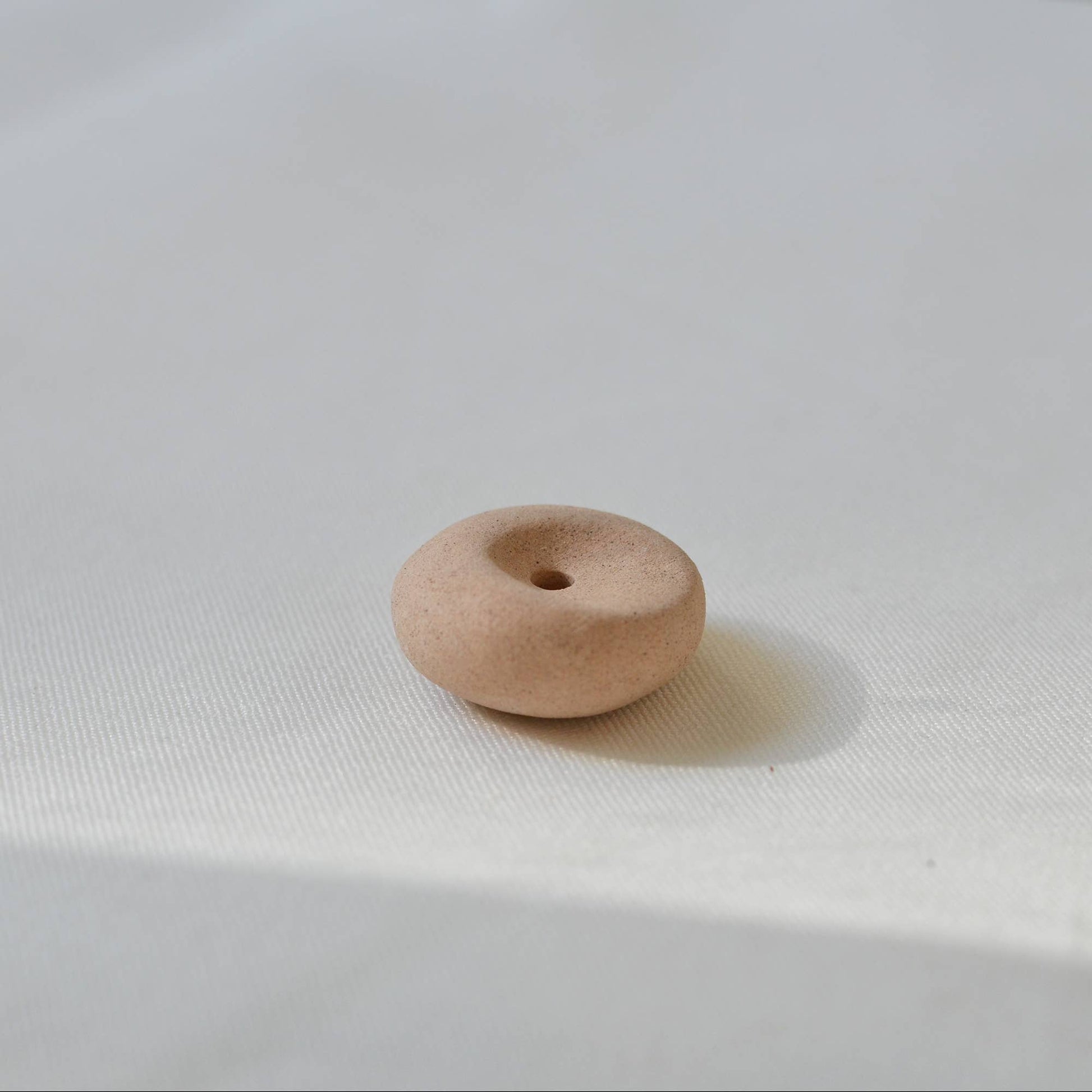 Clay pebble incense holder in soft beige tones, shown on a pale fabric background with a raw, organic finish.