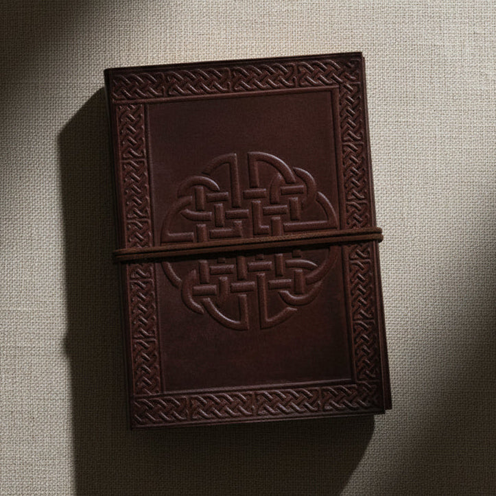 A brown embossed leather journal with a Celtic knot design on the cover, tied with a matching leather band.
