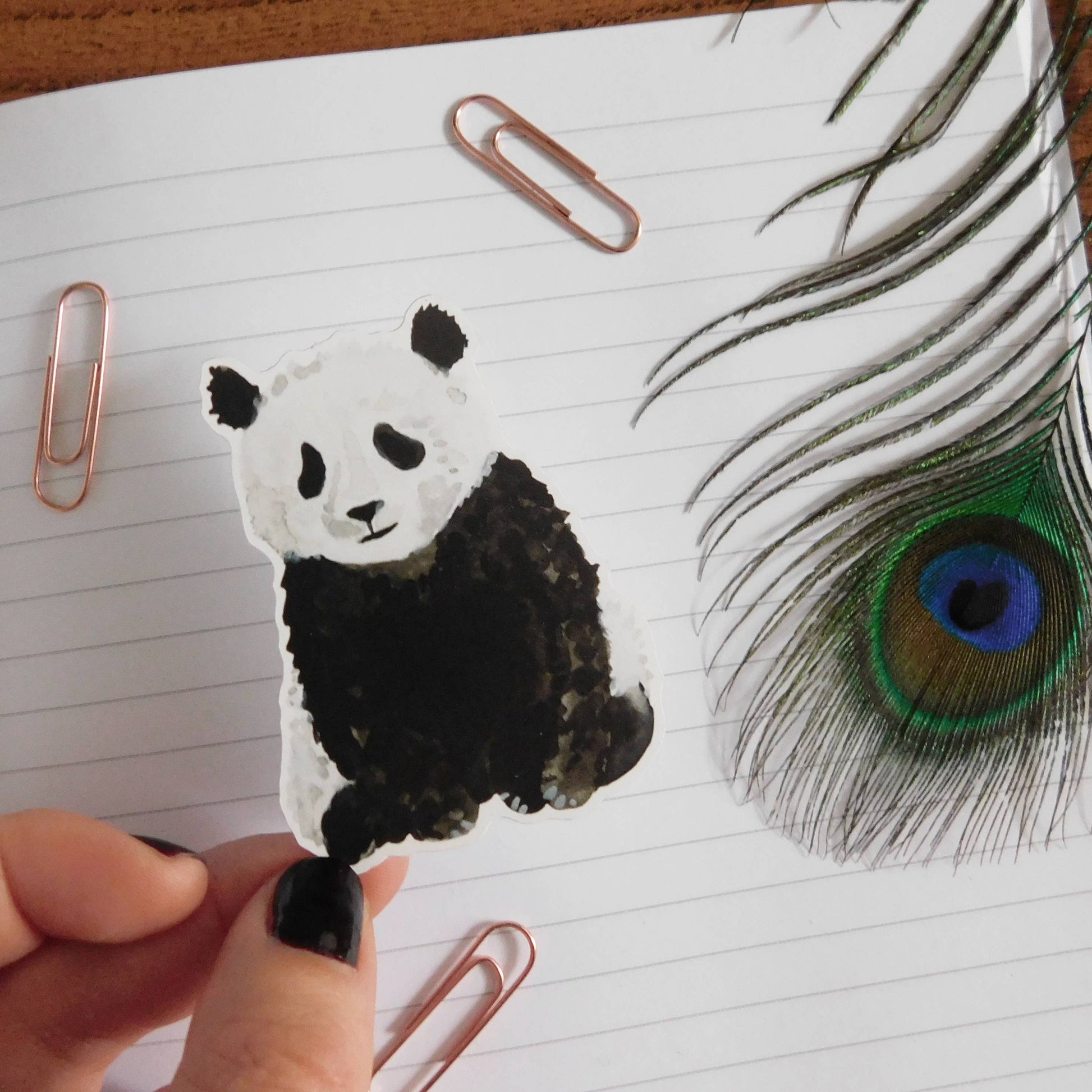 Hand holding a panda-shaped paper clip on lined paper with a peacock feather.