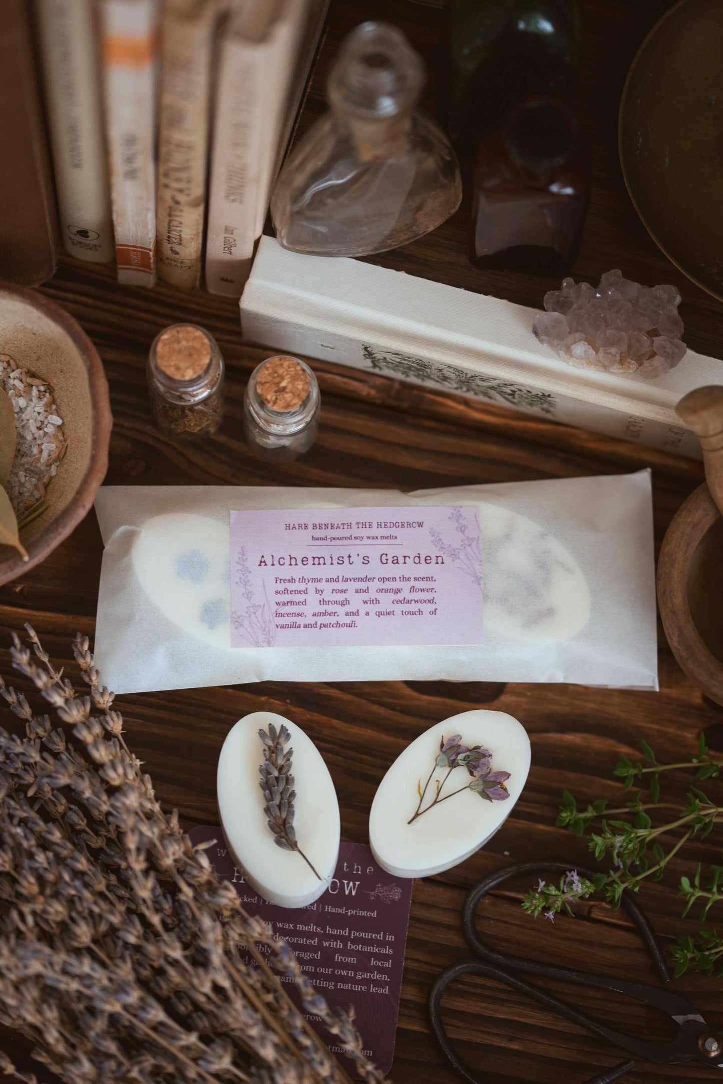 A styled flat-lay of handmade oval white wax melt with dried lavender sprigs, packaged in a translucent pouch labeled 'Alchemist's Garden'. The scene is decorated with books, crystals, small glass vials, and dried lavender on a dark wooden surface.