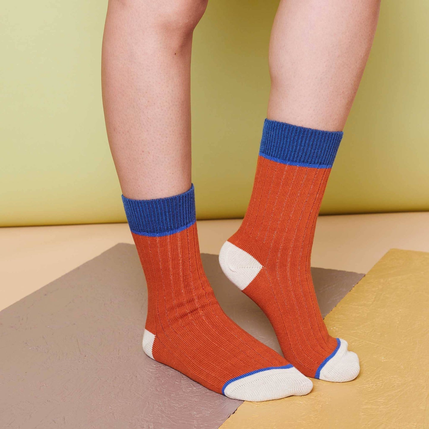 Orange organic cotton socks with blue cuffs and white toes worn by a person on a multicolored floor.