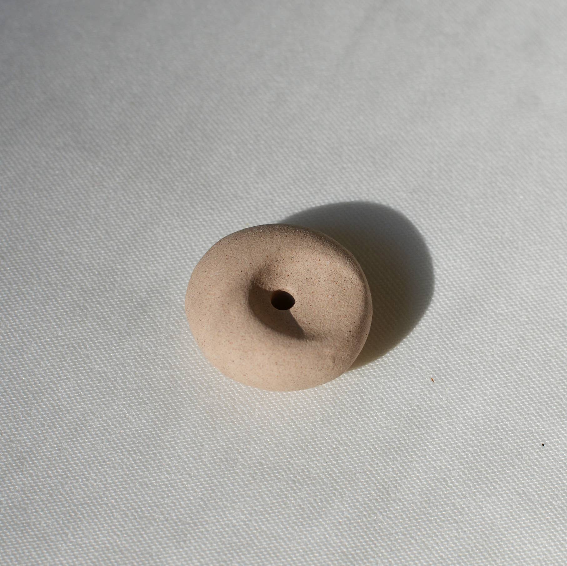 Clay pebble incense holder in soft beige tones, shown on a pale fabric background with a raw, organic finish.