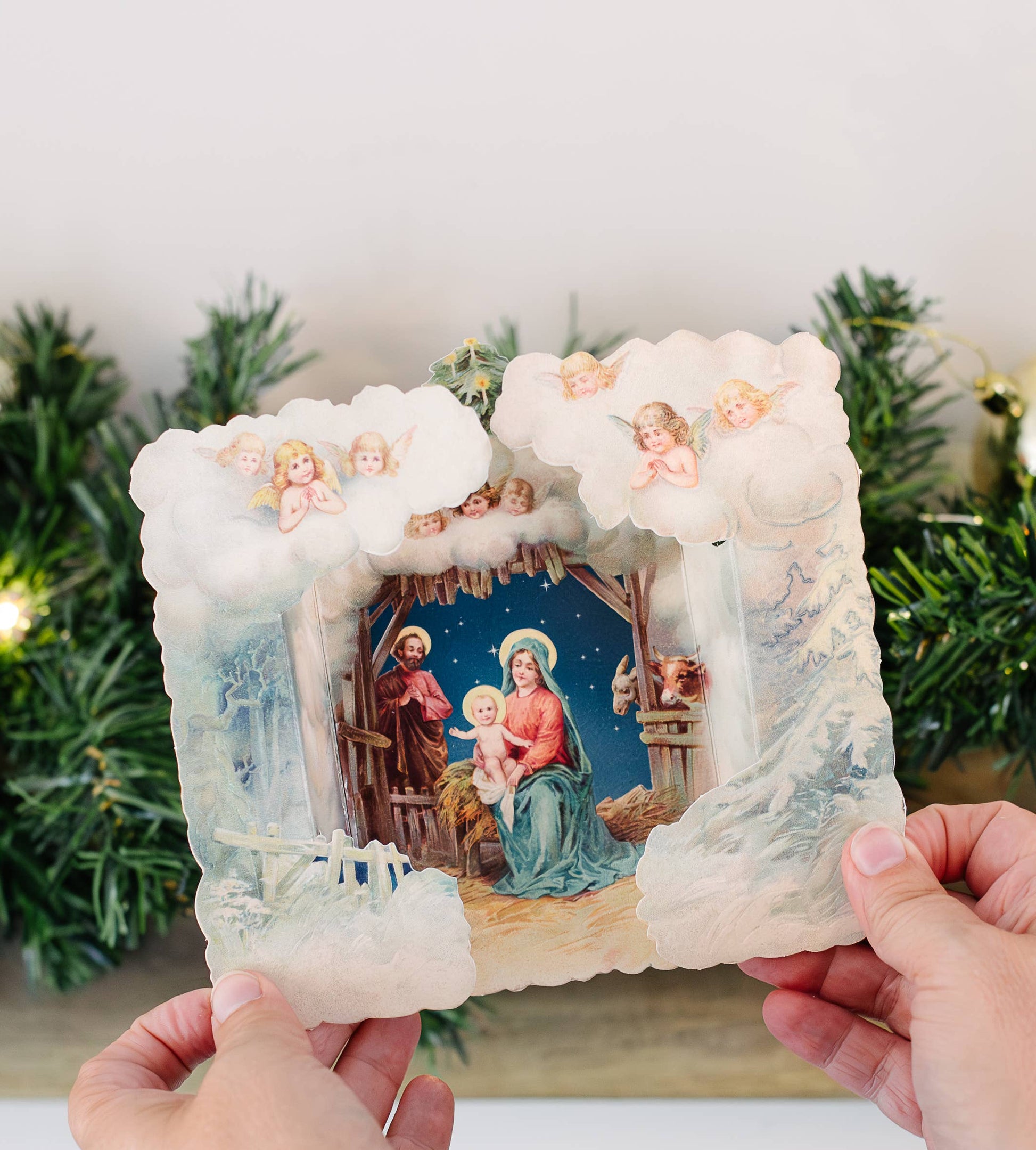 Decorative nativity scene card with a snow-covered surround held by hands against a festive background.